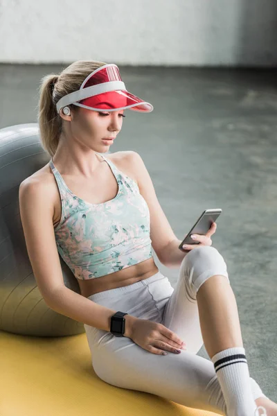 Schöne Sportlerin mit Smartwatch mit Smartphone in der Nähe von Fitnessball im Fitnessstudio — Stockfoto