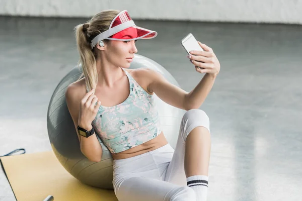 Веселая спортсменка со смартфоном, делающая селфи на смартфоне рядом с фитнес-мячом в спортзале — стоковое фото
