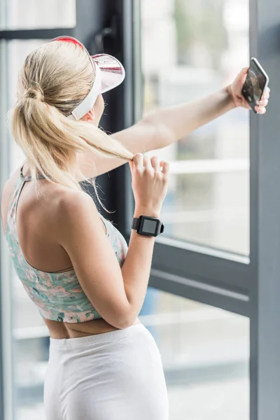 Rear view of sportswoman with smartwatch taking selfie on smartphone at gym — Stock Photo