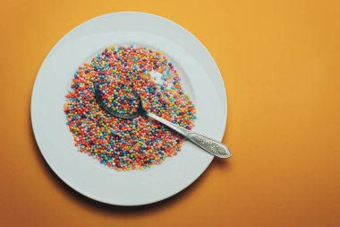 Turuncu zemin üzerinde vitaminler, boyalar, lezzet artırıcılar, besin takviyeleri, yenilikçi teknolojiler, şeker tatlılar bulunan beyaz tabak ve kaşık..