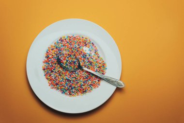 Turuncu zemin üzerinde vitaminler, boyalar, lezzet artırıcılar, besin takviyeleri, yenilikçi teknolojiler, şeker tatlılar bulunan beyaz tabak ve kaşık..