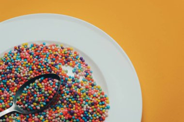 Turuncu zemin üzerinde vitaminler, boyalar, lezzet artırıcılar, besin takviyeleri, yenilikçi teknolojiler, şeker tatlılar bulunan beyaz tabak ve kaşık..