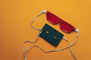 Turuncu arka planda kırmızı camlı, siyah manyetik kaset ve beyaz kulaklıklar.