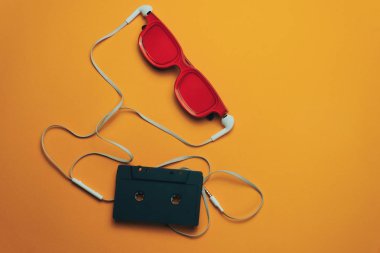 Turuncu arka planda kırmızı camlı, siyah manyetik kaset ve beyaz kulaklıklar.