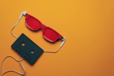 Turuncu arka planda kırmızı camlı, siyah manyetik kaset ve beyaz kulaklıklar.