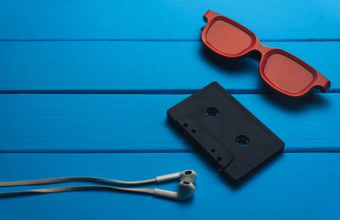 Kırmızı gözlük, klasik manyetik kaset ve mavi ahşap masa arka plan üzerinde beyaz kulaklıklar.