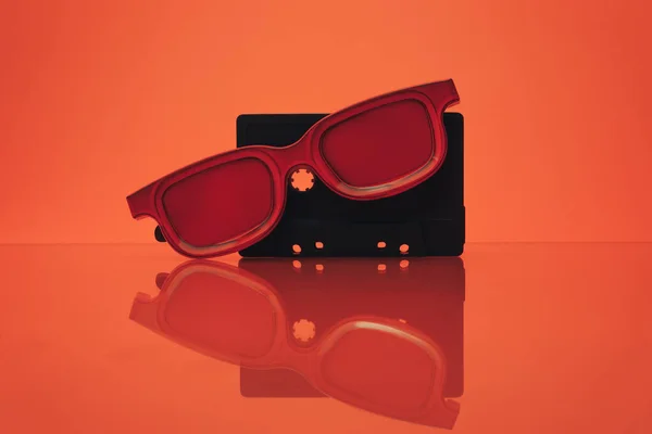 Black classic magnetic cassette tape and red glasses on a glass table. Beautiful coral orange background.