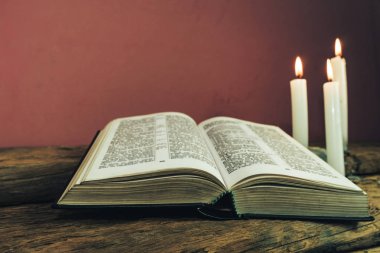 Yakın güzel açık İncil ve eski bir meşe ahşap masa ve kırmızı duvar arka plan üzerinde üç beyaz yanmış mumlar kadar. 