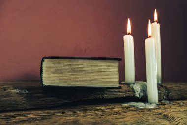 Güzel kapalı kutsal İncil ve eski bir meşe ahşap masa ve kırmızı duvar arka plan üzerinde üç beyaz yanmış mumlar. 