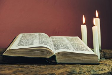 Yakın güzel açık İncil ve eski bir meşe ahşap masa ve kırmızı duvar arka plan üzerinde üç beyaz yanmış mumlar kadar. 