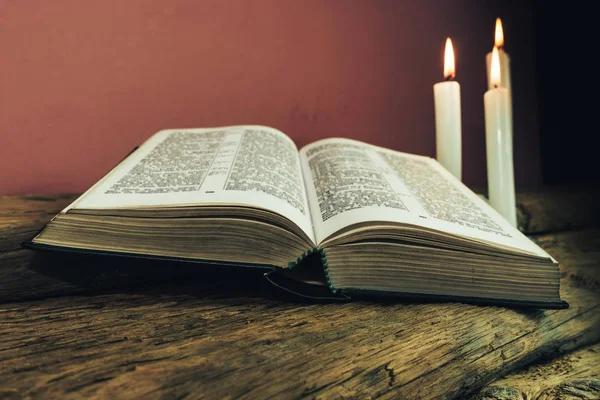 Close up beautiful open bible and l three white burned candles on a old oak wooden table and red wall background.