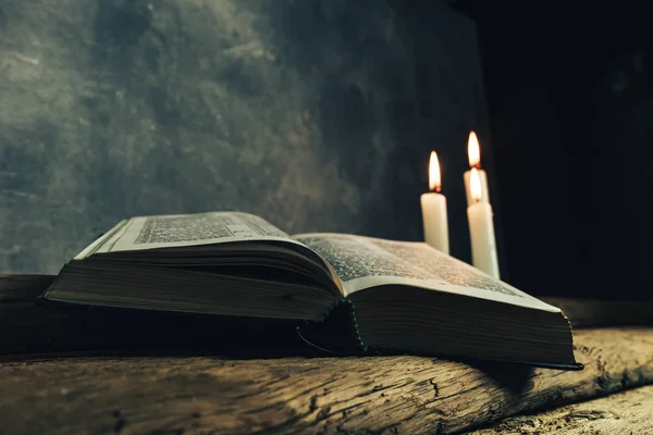 Beautiful open bible and three white burned candles on a old oak wooden table and dark gray wall background.