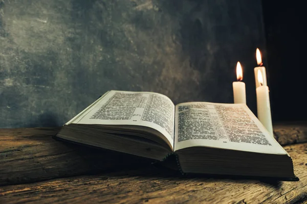 美しいオープン聖書と古いオーク材の木製のテーブルと暗い灰色の壁の背景に3つの白い焼かれたキャンドル — ストック写真