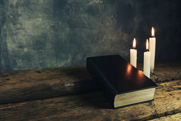 Güzel kapalı kutsal İncil ve eski bir meşe ahşap masa ve gri duvar arka plan üzerinde üç beyaz yanmış mumlar. 