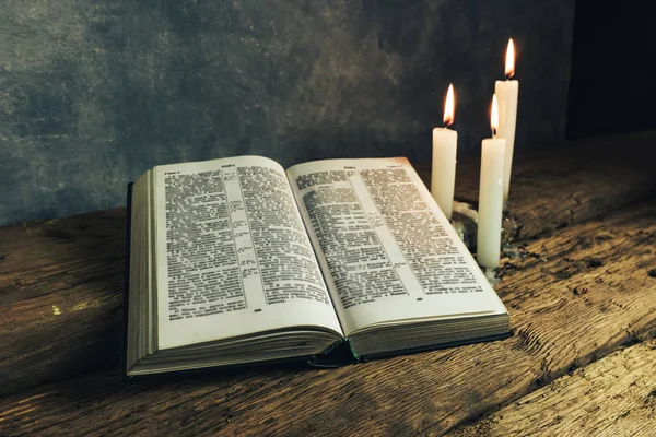 Güzel açık İncil ve eski bir meşe ahşap masa ve koyu gri duvar arka plan üzerinde üç beyaz yanmış mumlar. 