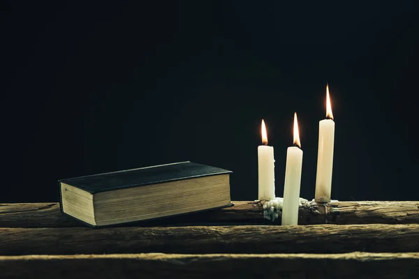 美丽的封闭的圣圣经和三个白色燃烧的蜡烛在一个古老的橡木表和黑色背景 — 图库照片