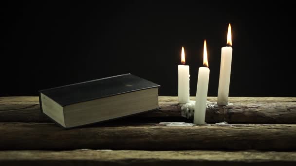 美しい閉鎖聖書と古いオーク材の木製のテーブルと黒い背景に3つの白い火のろうそく — ストック動画