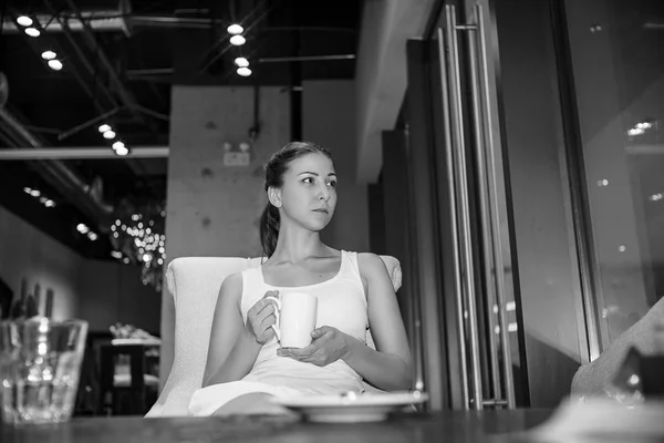 コーヒーの白いカップと白いドレスの美しい少女は 都市ビンテージ カフェとしんみりと遠く見る優しさに座っています 黒と白の写真 つ女性の屋内モデル ロイヤリティフリーのストック画像