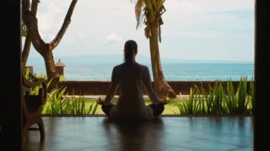 Kadın silüeti, mavi gökyüzü, Azure su, palmiye ağaçları ve yeşil çim ve ada Bali Endonezya, doğa sesleri ile güzel bir manzara ile okyanus plajında bungalovdan meditasyon, Lotus pozisyonunda oturan Yoga uygulayarak rahatlatıcı