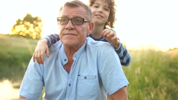 Un nieto rizado recurre a su abuelo y lo abraza por detrás. El abuelo le dice algo al niño y se ríe alegremente. felices vacaciones familiares — Vídeo de stock