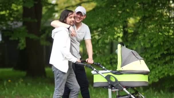 Um jovem pai e seu amigo estão andando com um carrinho no parque. Um casal homossexual com um bebé recém-nascido. Tolerância. Os homens abraçam, riem e mostram linguagem — Vídeo de Stock