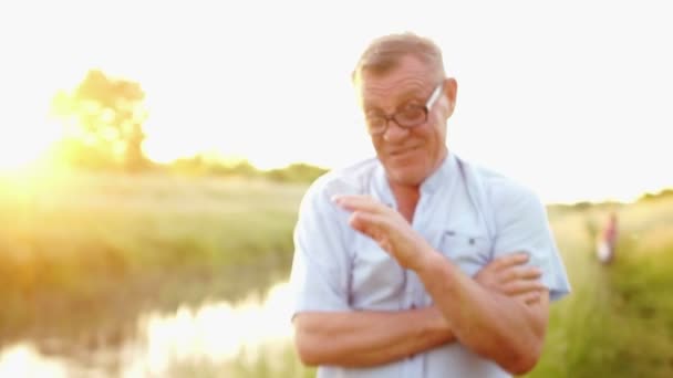 在河和夕阳的背景下, 有魅力的老人的肖像。替身, 动态视频。祖父挥舞着他的手, 看着相机。父亲日 — 图库视频影像