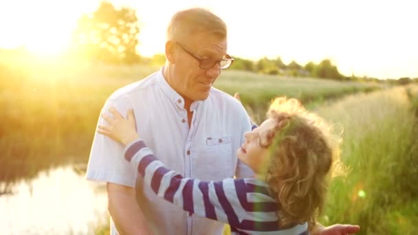Bejaarde opa en liefdevolle kleinzoon glimlachend en knuffelen elkaar buiten, aan de rivieroever. Twee generaties. Kleurrijke zonsondergang. Een gelukkige familie — Stockvideo