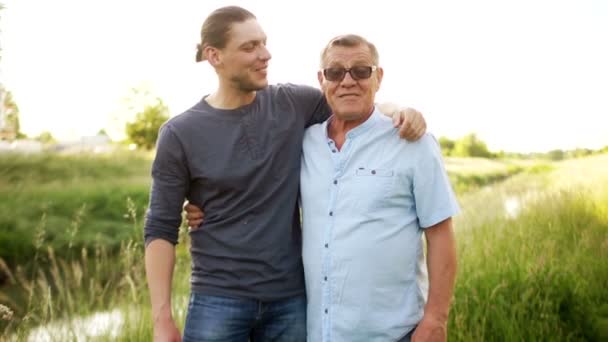 Filho adulto abraçando seu pai mais velho, na margem do rio. Duas gerações, conversa masculina, amizade masculina, dia dos pais. Idoso olha para a câmera — Vídeo de Stock