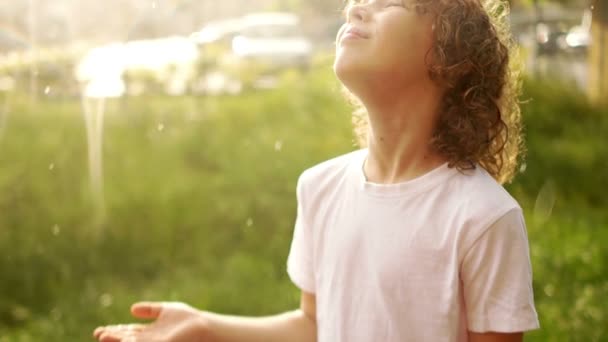 Een krullend schooljongen met natte haren staat onder de zomerregen en legt zijn hand in druppels. Zomerregen. Childrens dag — Stockvideo