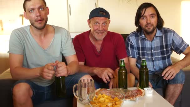 Los tres hombres miran fútbol y beben una cerveza. Un padre y dos de sus hijos adultos abren una cerveza. Fútbol fans — Vídeos de Stock