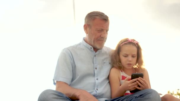 Зрелый мужчина и маленькая девочка кудрявая школьница смотрят видео в смартфоне. На фоне голубого неба. День Отцов. День семьи — стоковое видео
