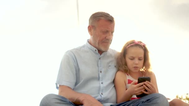 Mogen man och lilla flicka curly Skolflicka titta på videon i smartphone. Mot den blå himlen. Fars dag. Familjedag — Stockvideo