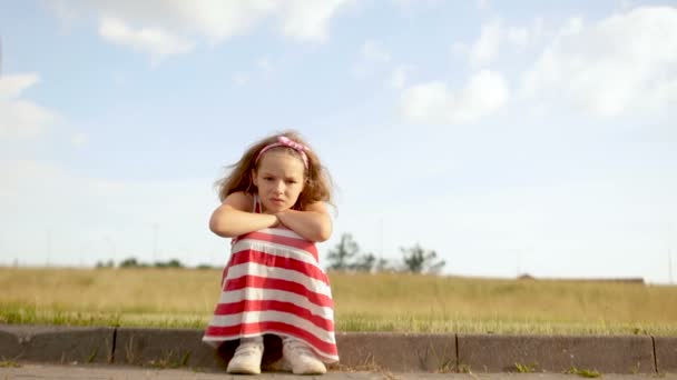 Самотня кучерява дівчина сидить на краю дороги проти блакитного неба. Дівчина сумна і сердита — стокове відео