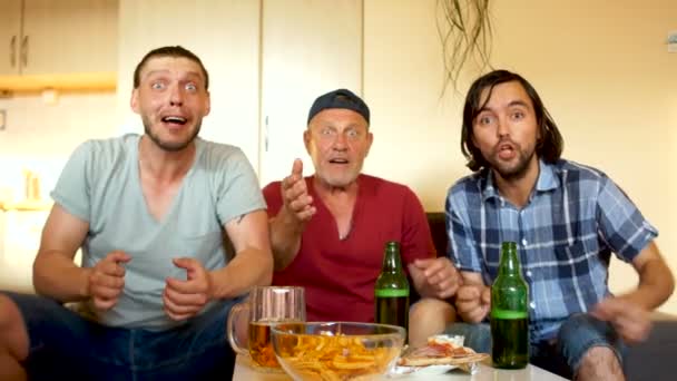 Los hombres ven un partido de fútbol en la televisión. Tres hombres beben cerveza y comen bocadillos, sentados en un sofá frente a una mesa de café. Abanicos fieles, fanáticos del bootball — Vídeo de stock