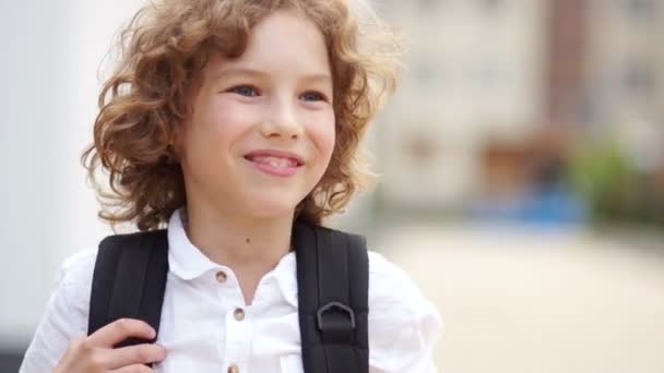 O menino de olhos azuis corrige os arreios da mochila em seu ombro e joga para trás cabelos encaracolados. Close up retrato, de volta à escola, menino feliz da escola — Vídeo de Stock