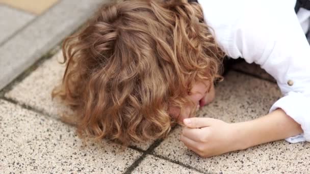 Kıvırcık okul çocuğu eğleniyor ve dalga geçmeyi. Çocuk asfalt üzerine düştü ve yumruğunu kaldırım taşı üzerinde darbelere. Tüm Zafer Bayramı, şaka, eğlenceli gülmek, okula dönüş — Stok video