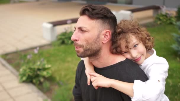 Un joven con una cara seria se sienta en un banco. es abrazado por un chico de pelo rizado. Padre e hijo, día del padre — Vídeos de Stock