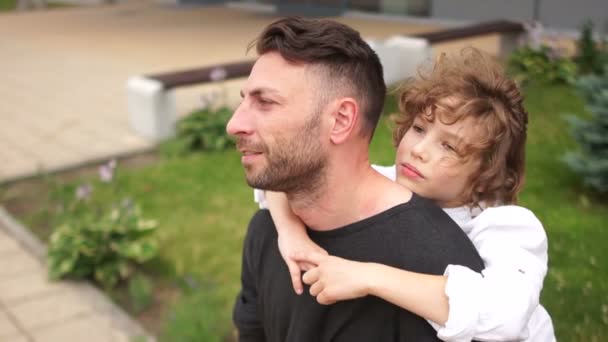 Hombre elegante y rizado adolescente niño sentado en un banco. El viento sopla y mueve su cabello. Miran pensativamente a la distancia. Padre e hijo — Vídeo de stock