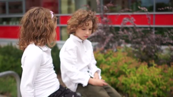 Um menino e uma menina estão sentados ao lado deles no corrimão perto da escola e olhando um para o outro. Amizade escolar, primeiro amor — Vídeo de Stock