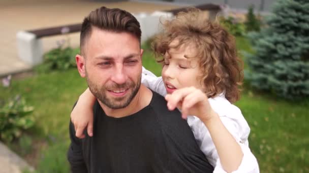 Hijo y padre hablando, sentados en un banco del parque. El chico gesticula animadamente y se ríe. Un padre joven, una familia feliz, una familia monoparental — Vídeos de Stock