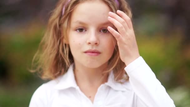 Um retrato próximo de uma menina com cabelo encaracolado e olhos castanhos. A criança está vestida com uma camisa branca. Endireita o cabelo com a mão, olha a sério, calmo e pensativo — Vídeo de Stock