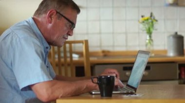 Yaşlı bir adam olarak sosyal ağlar bir dizüstü bilgisayar kullanarak iletişim kurar. Modern teknoloji, yaşlılar günü. Bir adam mutfak ve içme çay veya kahve içinde oturuyor
