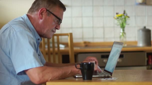 一位老人用手提电脑在社交网络上交流。现代科技, 老人之日。一个男人坐在厨房里喝茶或喝咖啡 — 图库视频影像