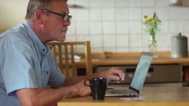 Compras en línea. Un pensionista bebe té en la cocina y paga sus facturas a través de Internet. Tecnologías modernas, personas mayores, jubilación tranquila, edad de jubilación. Vestidos gafas para ver mejor — Vídeo de stock