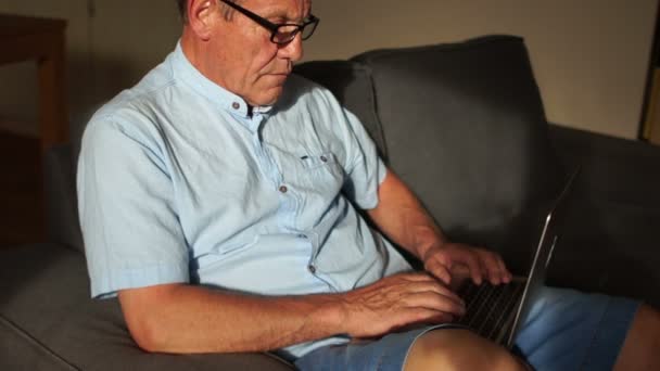 Ein reifer Mann arbeitet mit einem Noutbeck, der auf der Couch sitzt. Fernbeschäftigung, Internet-Banking für Rentner — Stockvideo