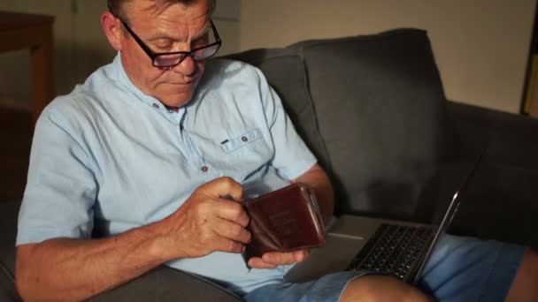 Nyugdíjas fizeti a számlákat egy internetes és egy laptop segítségével. a férfi előveszi a bankkártyával a tárcáját. Ő ül a kanapén, ő egy laptop ölében — Stock videók