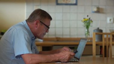 Emekli insanlar için Internet teknolojisi. Yaşlı bir adam diz üstü ile mutfak masasında otururken çalışıyor. Gözlüklerini yükseltir ve bakmak daha iyi olur ekran doğru eğilir