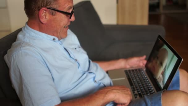 Senior man speaks in skype with his son and small grandson. He waves to them. A happy family — Stock Video