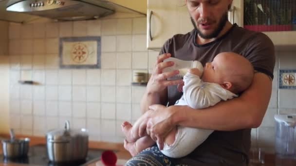 Moderna unga far matar ett barn från en flaska i köket. En kastrull soppa kokar på spisen. När mamma inte är hemma. Ensamstående far, fars dag — Stockvideo