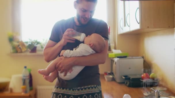 A young hipster father feeds from a baby bottle in the kitchen. The man smiles at his son and talks to him kindly. Fathers Day — Stock Video
